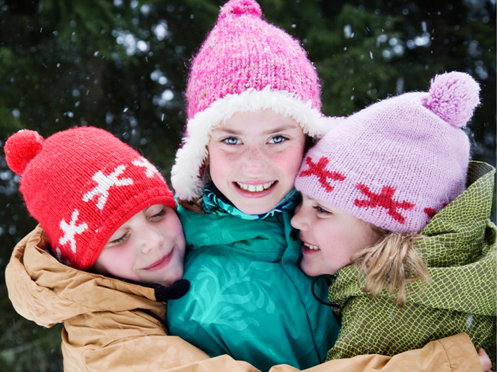 Three children wearing colorful winter hats smile and hug each other outdoors, surrounded by snow and trees.